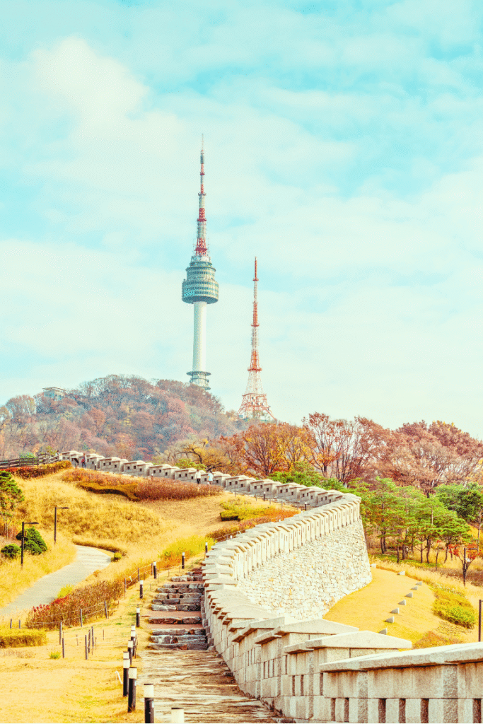 alt="南山公園の城郭とNソウルタワーを望む風景。秋色に染まった木々と曲線を描く石垣が印象的。"


